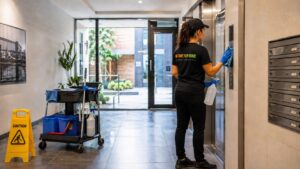 NTFG cleaner wiping lift buttons in a modern strata apartment lobby with cleaning trolley nearby.